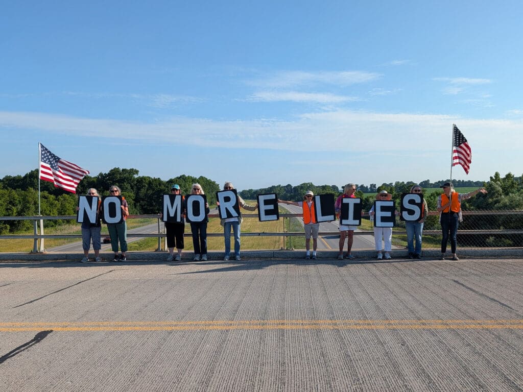 Protesters holding No More Lies signs on bridge with American flags