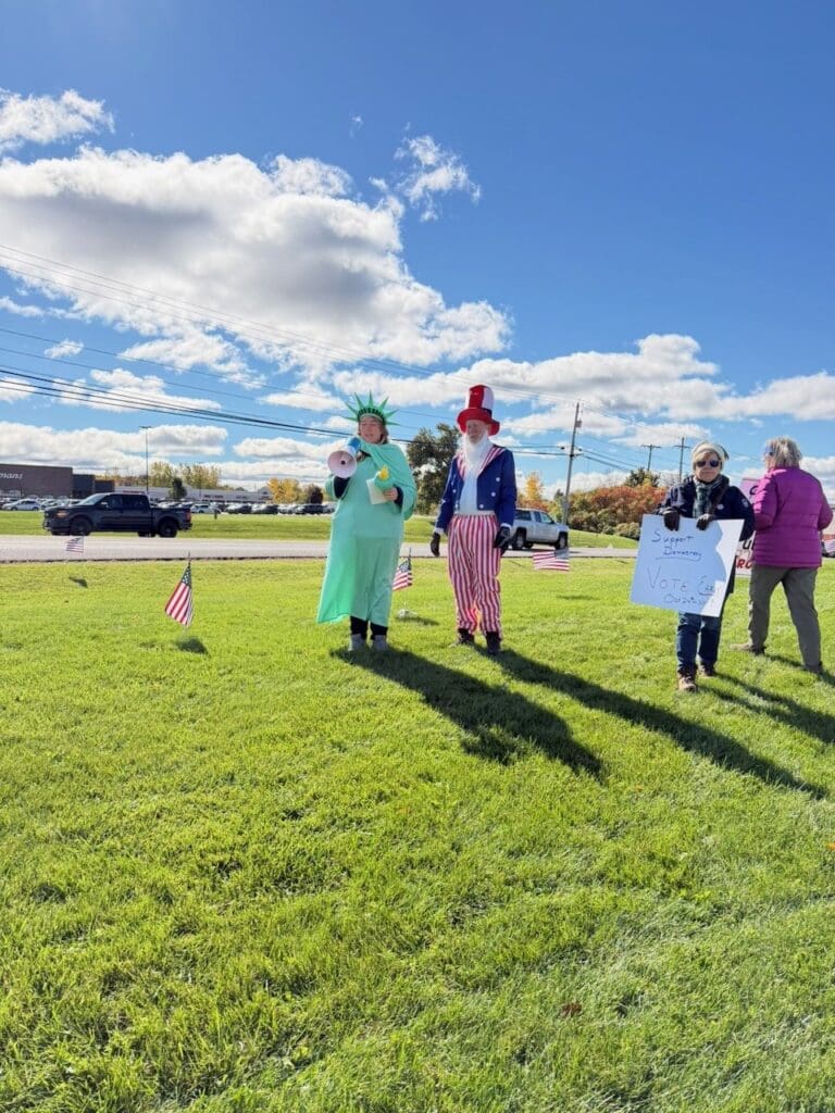 Volunteers dressed as the Statue of Liberty, holding a megaphone, and Uncle Sam stand on the grass beside a woman holding a "Support Democracy, Vote Early" sign