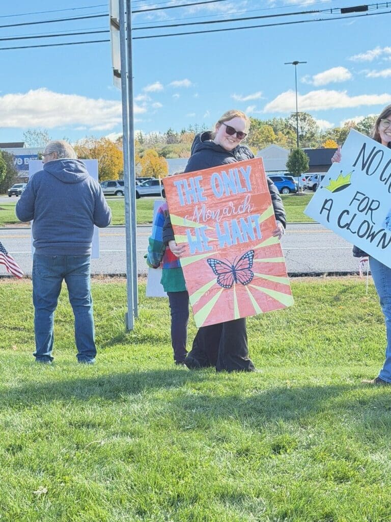 Smiling woman holding a sign with a monarch butterfly that reads "The only Monarch we want"