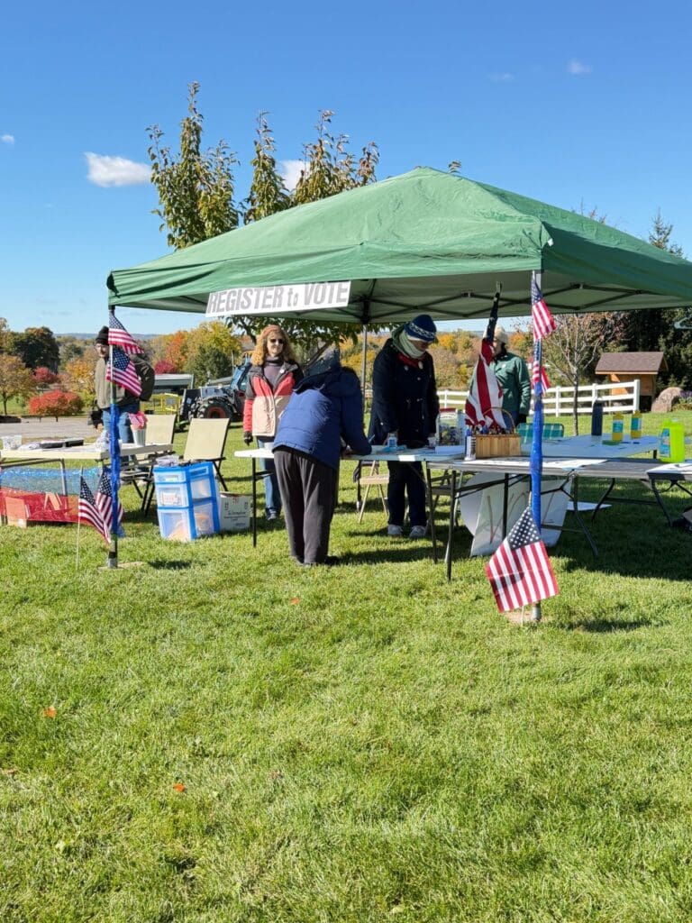 Volunteers help a visitor at a table under a green "Register to Vote" tent decorated with American flags