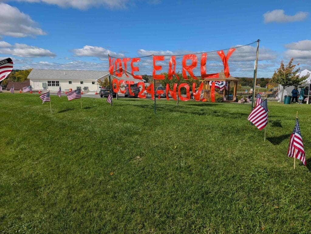 Large orange letters on netting read "Vote Early Oct. 24 - Nov. 1" above a lawn lined with small American flags